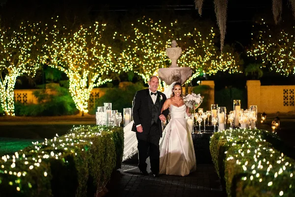A bride and groom walking through a garden path decorated with string lights and candles at a wedding venue.