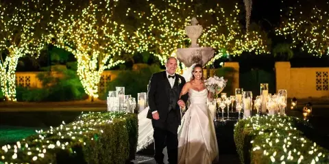 A bride and groom walking through a garden path decorated with string lights and candles at a wedding venue.