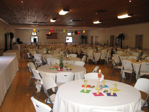 Interior of a large community hall set up for an event with round tables and white chairs.