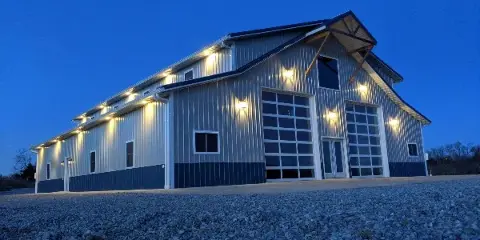 Exterior view of a large metal barn event venue with illuminated exterior lights at dusk.