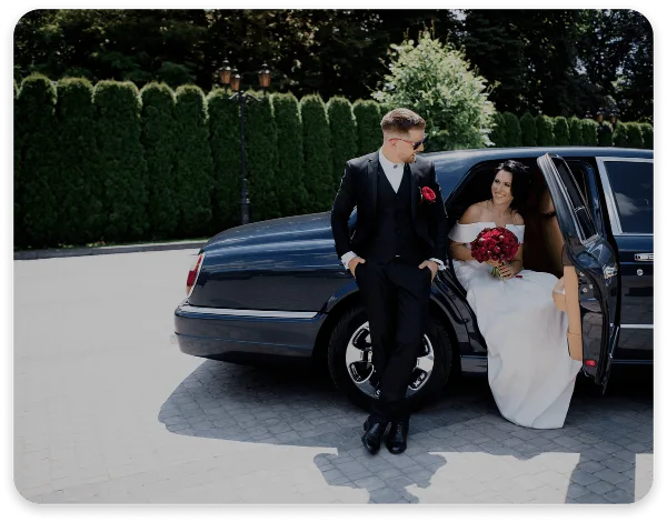 A bride and groom posing next to a black luxury sedan with the door open.