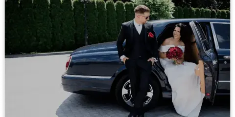A bride and groom posing next to a black luxury sedan with the door open.