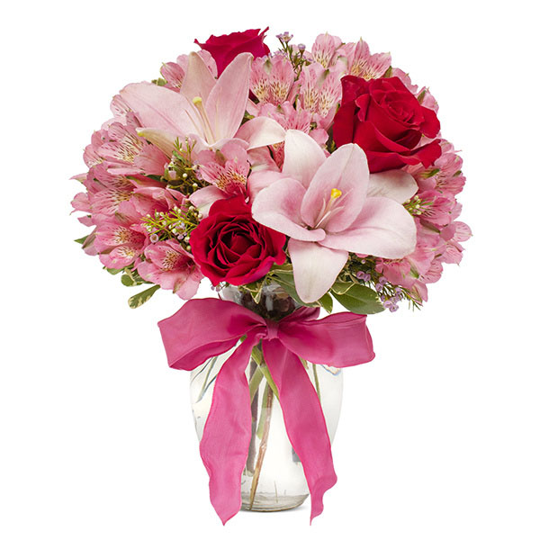 A hand-arranged floral bouquet containing red roses, pink lilies, and alstroemeria in a clear glass vase with a pink ribbon.