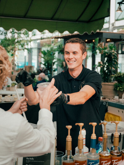 A smiling catering staff member in a black polo shirt serves a drink to a guest at an event, with beverage dispensers in the foreground.