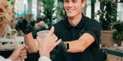 A smiling catering staff member in a black polo shirt serves a drink to a guest at an event, with beverage dispensers in the foreground.