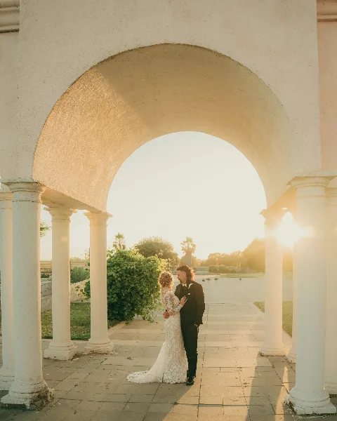A bride and groom standing together under a white stone archway during a wedding photoshoot.