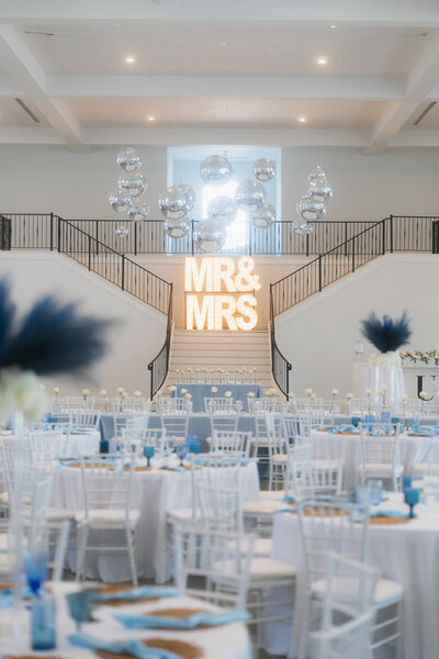 A wedding reception venue with white Chiavari chairs, round tables with blue and gold accents, and a large illuminated 'MR & MRS' sign on a staircase.