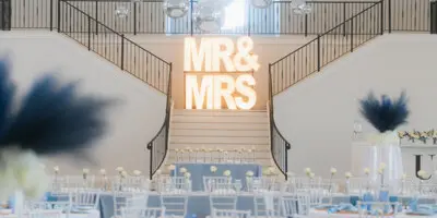 A wedding reception venue with white Chiavari chairs, round tables with blue and gold accents, and a large illuminated 'MR & MRS' sign on a staircase.