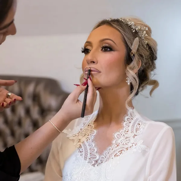 A makeup artist applying lip liner to a bride wearing a white lace dress and hair accessories.