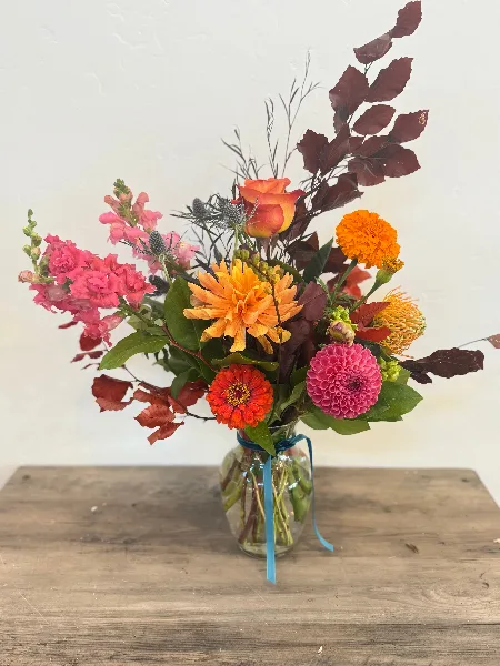 A colorful floral arrangement featuring dahlias, roses, and foliage in a glass vase on a wooden table.