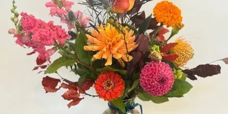 A colorful floral arrangement featuring dahlias, roses, and foliage in a glass vase on a wooden table.