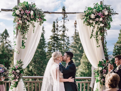 A wedding couple kissing under a floral arch decorated with white and pink flowers and white drapery.