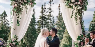 A wedding couple kissing under a floral arch decorated with white and pink flowers and white drapery.