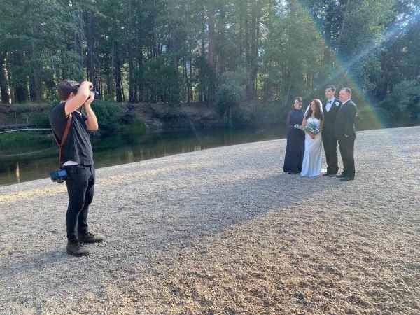A wedding couple and officiant standing outdoors near a wooded creek while a photographer takes pictures.