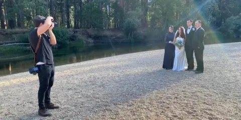A wedding couple and officiant standing outdoors near a wooded creek while a photographer takes pictures.