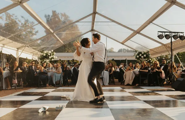 A couple dances on a checkered floor inside a large clear-frame tent at a wedding reception.