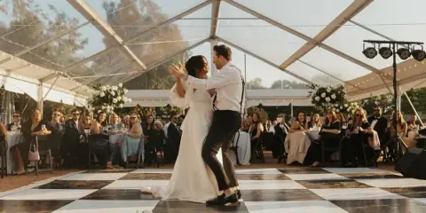 A couple dances on a checkered floor inside a large clear-frame tent at a wedding reception.