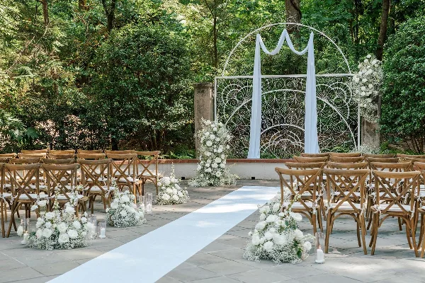 Outdoor wedding ceremony setup with a white metal arch, floral arrangements, and wooden chairs.