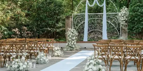Outdoor wedding ceremony setup with a white metal arch, floral arrangements, and wooden chairs.