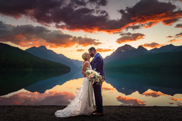 A bride and groom kissing in front of a mountain lake at sunset
