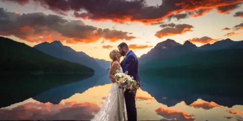 A bride and groom kissing in front of a mountain lake at sunset