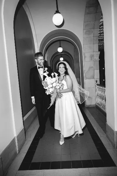 A black and white photograph of a bride and groom walking through an arched hallway, with the bride holding a bouquet.