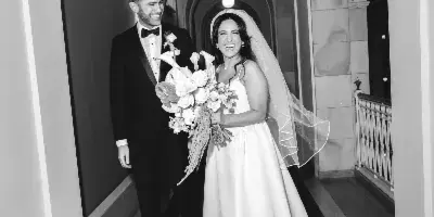 A black and white photograph of a bride and groom walking through an arched hallway, with the bride holding a bouquet.