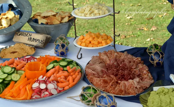 An outdoor catering table featuring a large vegetable platter, a charcuterie board, hummus, and a tiered cheese display.