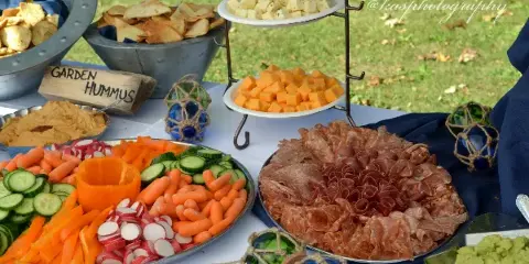 An outdoor catering table featuring a large vegetable platter, a charcuterie board, hummus, and a tiered cheese display.