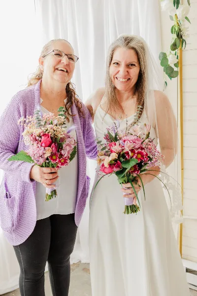 Two women, one in a wedding dress and veil, standing together holding floral bouquets in a bright indoor ceremony space.