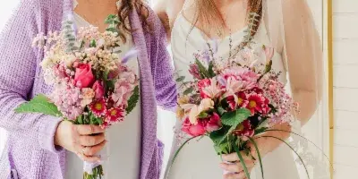 Two women, one in a wedding dress and veil, standing together holding floral bouquets in a bright indoor ceremony space.