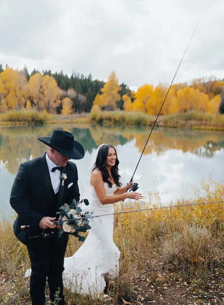A bride and groom in formal wedding attire holding fishing rods by a lake with autumn foliage in the background.