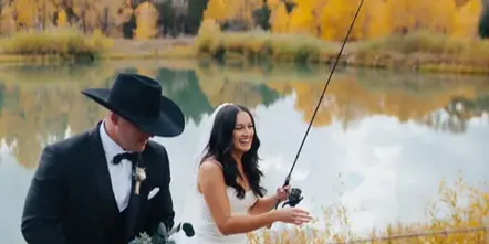 A bride and groom in formal wedding attire holding fishing rods by a lake with autumn foliage in the background.
