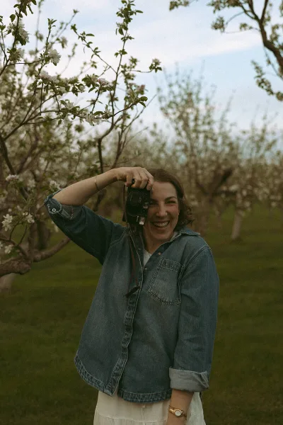 A smiling woman holding a camera in a blooming orchard