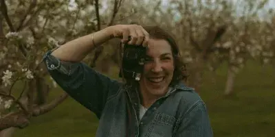 A smiling woman holding a camera in a blooming orchard