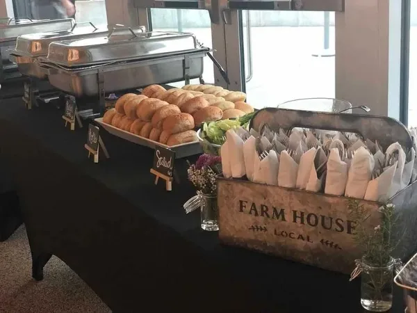 A catering table featuring chafing dishes, a tray of bread rolls, and a rustic wooden bin labeled 'FARM HOUSE LOCAL' holding wrapped cutlery.