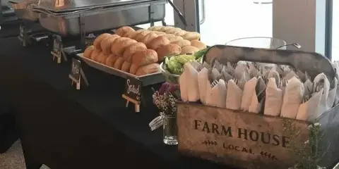 A catering table featuring chafing dishes, a tray of bread rolls, and a rustic wooden bin labeled 'FARM HOUSE LOCAL' holding wrapped cutlery.