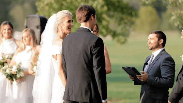 A bride and groom standing at an outdoor wedding ceremony with an officiant holding a tablet.