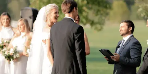 A bride and groom standing at an outdoor wedding ceremony with an officiant holding a tablet.