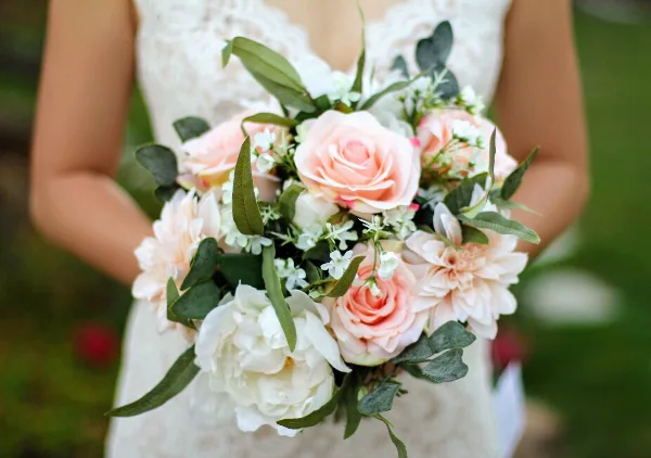 A bride in a white lace dress holding a bouquet of pink roses, white peonies, and greenery.