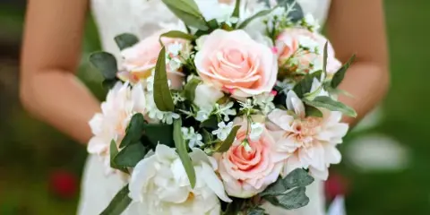 A bride in a white lace dress holding a bouquet of pink roses, white peonies, and greenery.