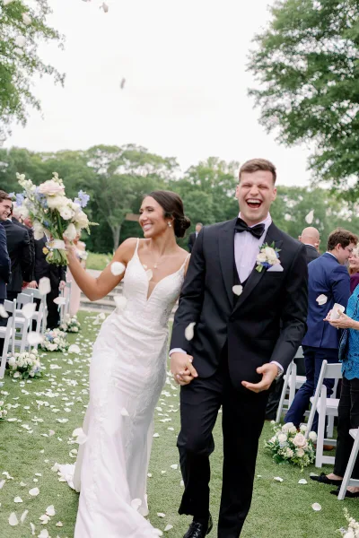 A bride and groom walking down an outdoor wedding aisle with white chairs and floral decorations.