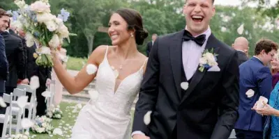 A bride and groom walking down an outdoor wedding aisle with white chairs and floral decorations.