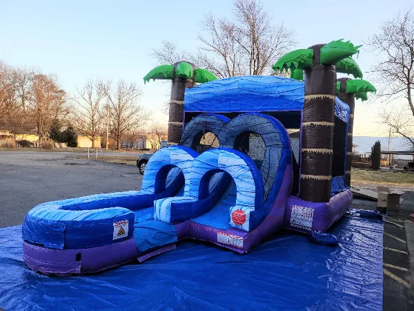 A blue and purple inflatable bounce house with a slide and palm tree accents set up outdoors on a blue tarp.