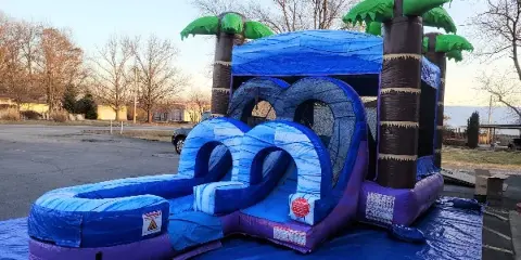 A blue and purple inflatable bounce house with a slide and palm tree accents set up outdoors on a blue tarp.