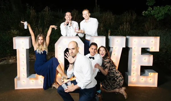 A group of wedding guests posing playfully with large, illuminated marquee letters spelling 'LOVE' at night.