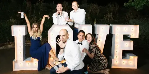 A group of wedding guests posing playfully with large, illuminated marquee letters spelling 'LOVE' at night.