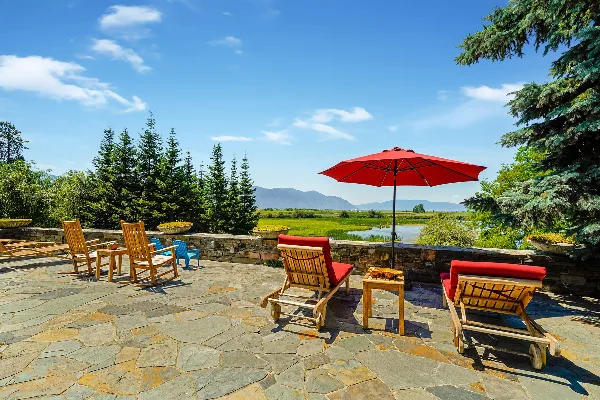 Outdoor stone patio with wooden lounge chairs and a red umbrella overlooking a lake and mountains.