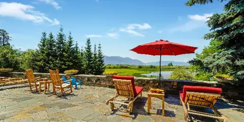 Outdoor stone patio with wooden lounge chairs and a red umbrella overlooking a lake and mountains.