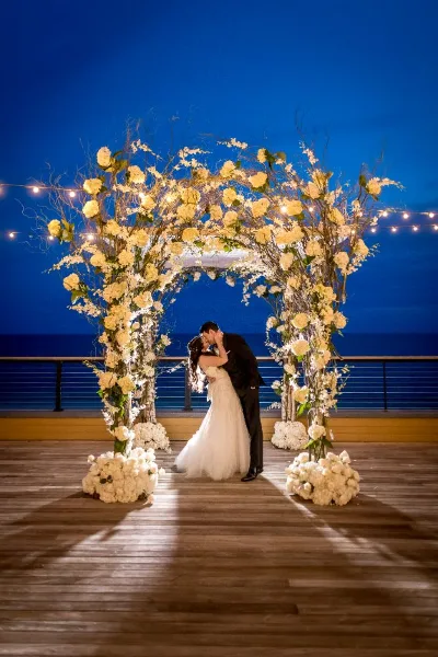 A wedding couple kissing under a large floral arch decorated with white roses and branches, set against a dark blue evening sky.